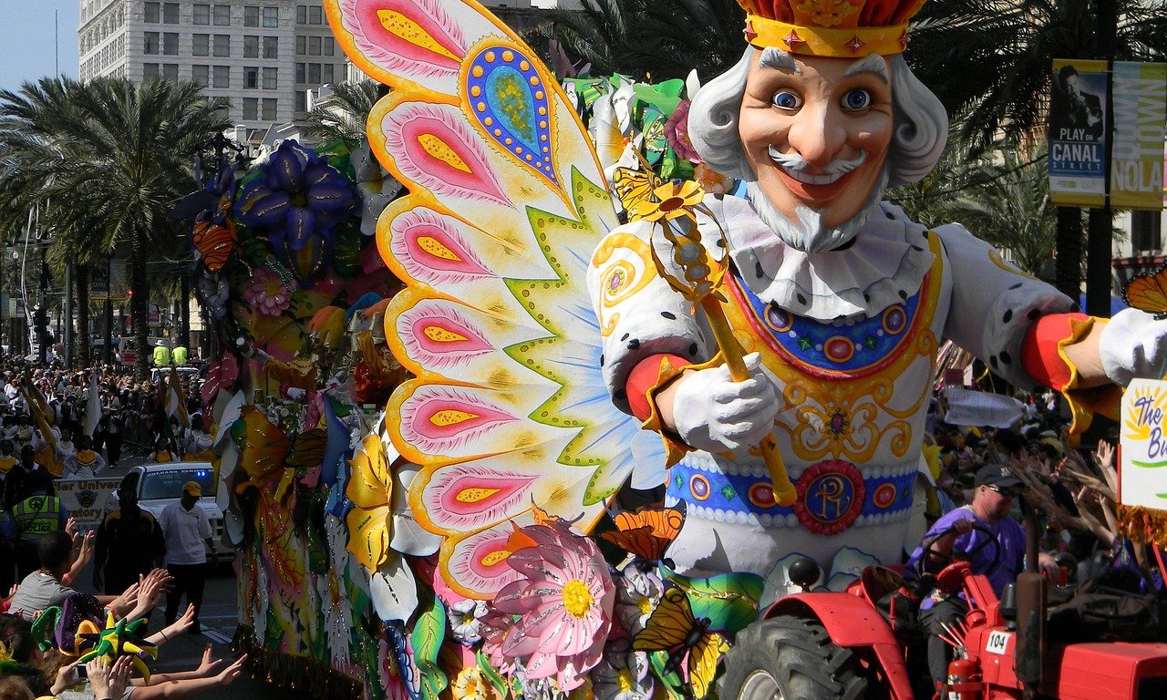 Ein reich dekorierter Mardi Gras Wagen mit einem weißhaarigen, geflügelten König als Galleonsfigur. Im Hintergrund sind Menschen in bunten Kostümen zu sehen, die Ketten mit bunten Perlen in die Menge werfen.