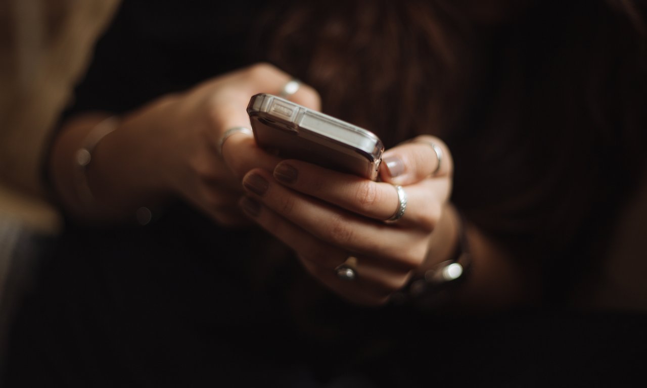 Eine Frau hält ein Mobiltelefon in Händen, der Hintergrund ist dunkel, man sieht auch nur die Hände und das Telefon.