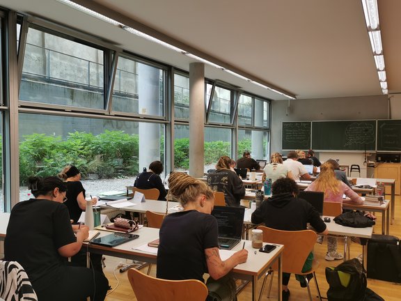 Studierende arbeiten an gereihten Tischen an ihren Schreibprojekten