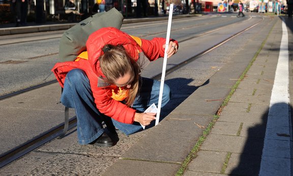 Eine Frau ist in Hockstelle neben dem Gehsteig und hat einen Zollstock in der Hand.