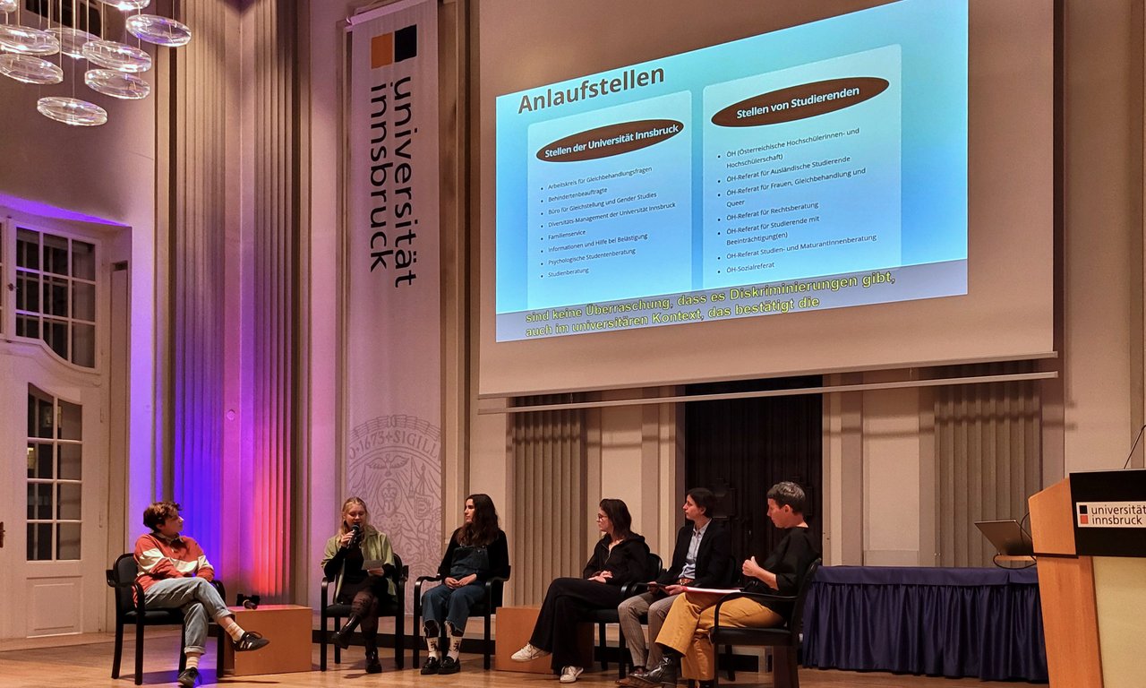 Mehrere Personen sitzen auf einem Podium und diskutieren, im Hintergrund eine Leinwand.