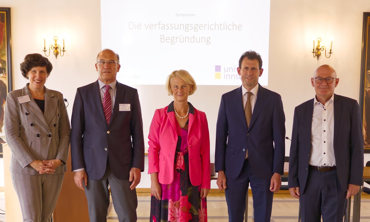 Fünf Personen stehen für ein Gruppenfoto vor einer Leinwand, auf der „Die verfassungsgerichtliche Begründung“ zu lesen ist.