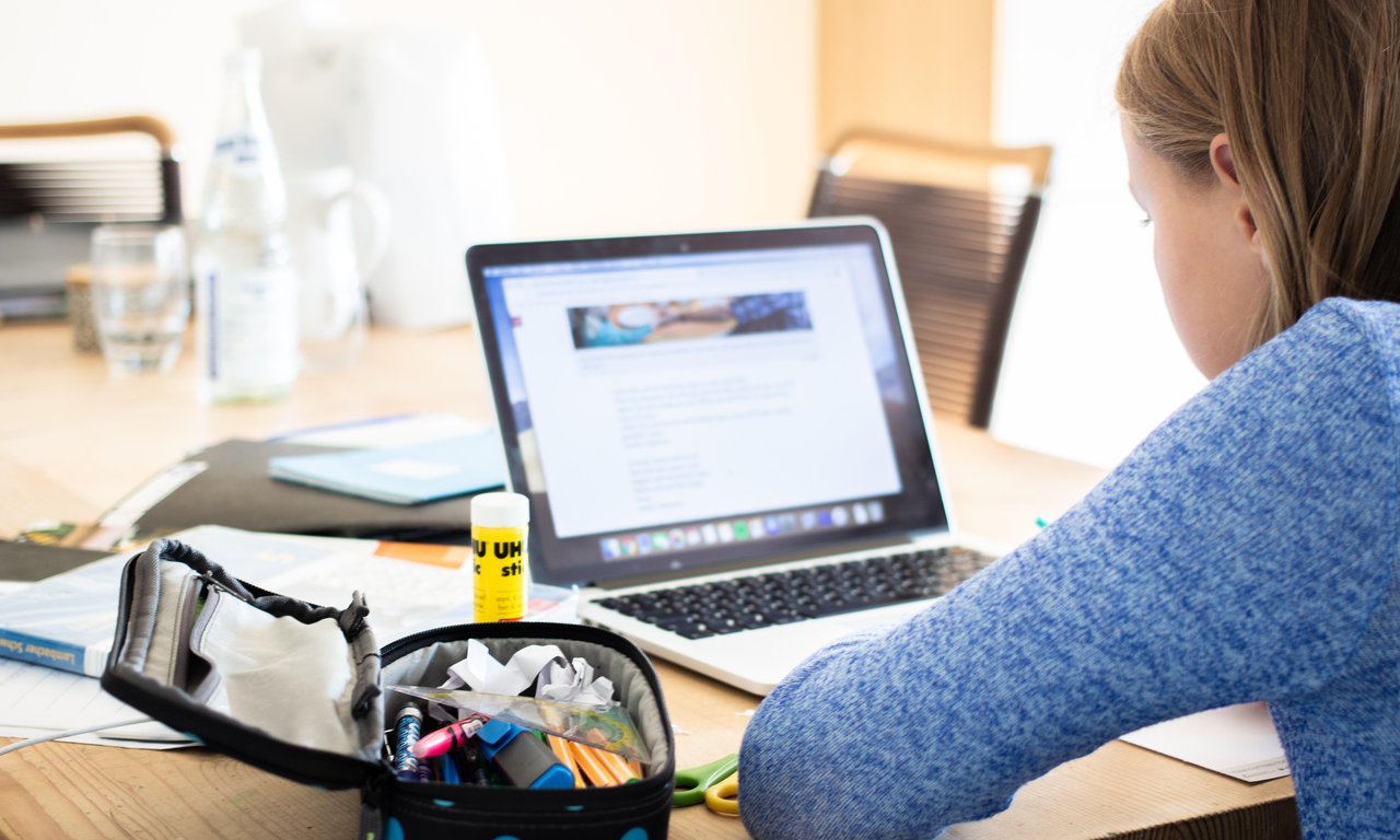 Mädchen sitzt vor einem Laptop, man sieht den Bildschirm, das Mädchen nur von der Seite. Außerdem sind Schulutensilien im Bild zu sehen.