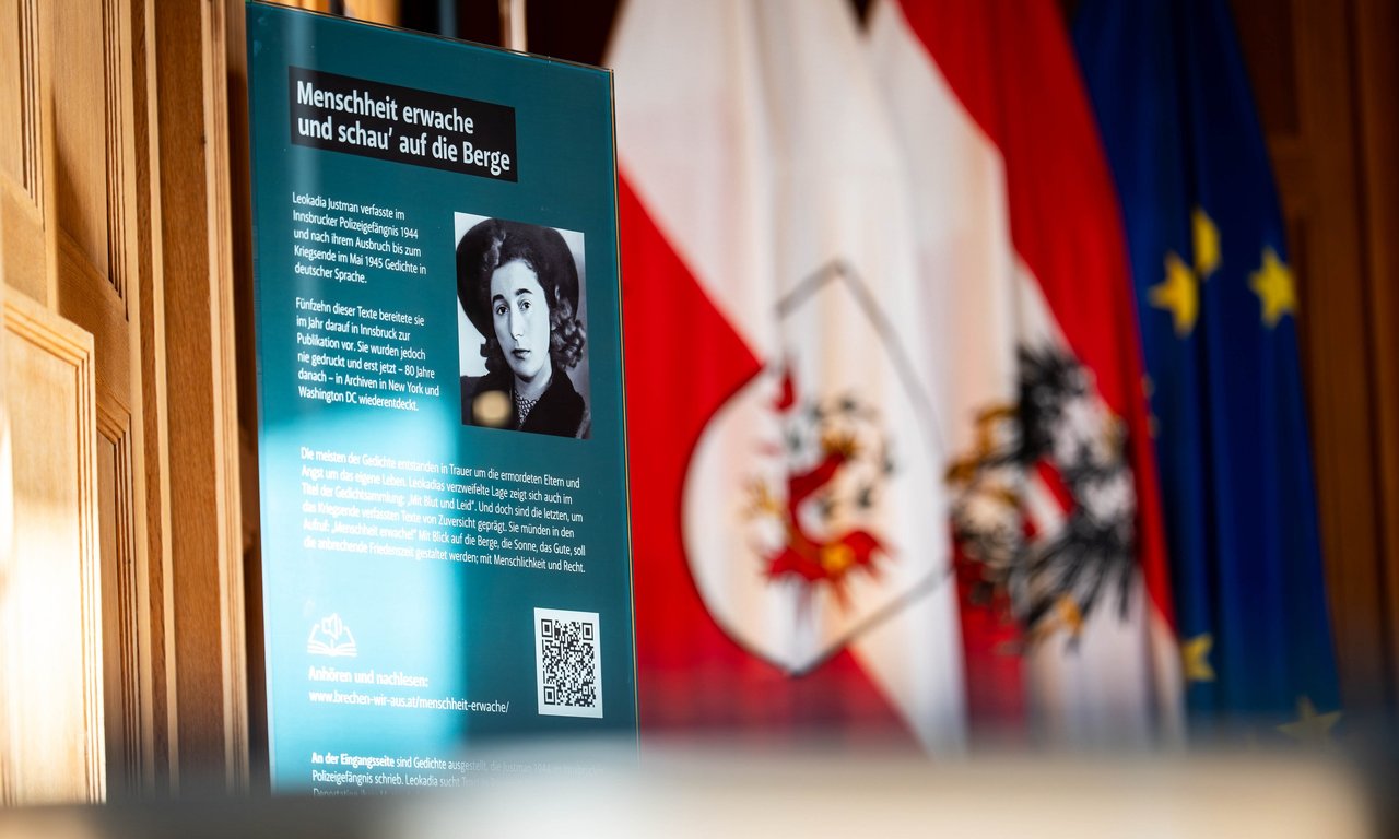 Ausstellungstafel mit schwarzweißem Bild von Leokadia Justmann und Infotext sowie QR Code zu Audiofassung der Gedichte; Tiroler Landesflagge im Hintergrund