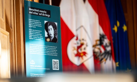 Ausstellungstafel mit schwarzweißem Bild von Leokadia Justmann und Infotext sowie QR Code zu Audiofassung der Gedichte; Tiroler Landesflagge im Hintergrund