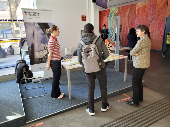 Zu sehen sind junge Erwachsene. Sie stehen vor dem Informationsstand der Katholisch-Theologischen Fakultät.