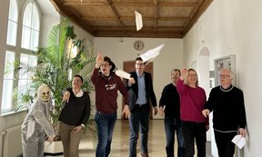 Zu sehen sind die Mitglieder des Instituts für Praktische Theologie. Einige von ihnen tragen ein Faschingskostüm. Sie werfen Papierflieger.