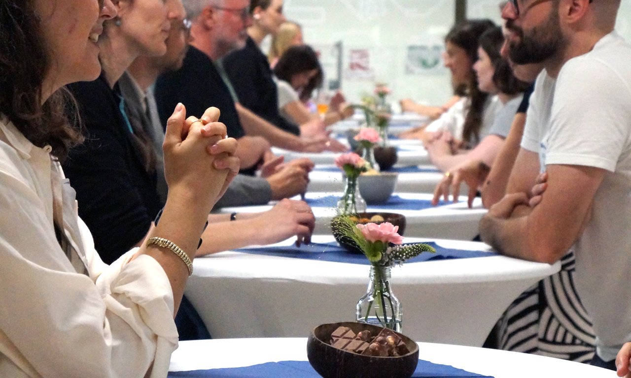 Menschen stehen einander an Stehtischen gegenüber, jeweils zwei, und reden miteinander. Sichtbar sind sieben solche Paarungen, die Tische sind mit einer weißen Tischdecke und Blumen geschmückt.