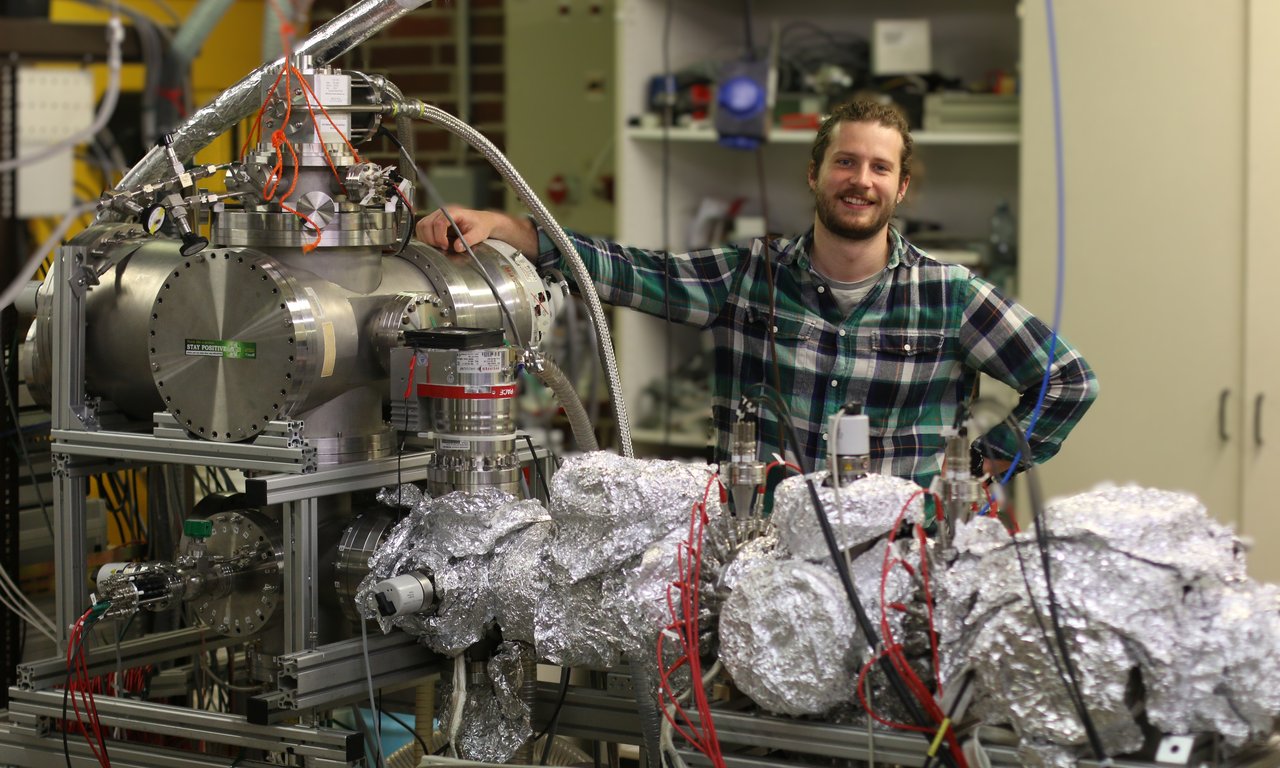 Young man in a plaid shirt in a laboratory setting stands next to a large, metallic experimental setup. Many pipes, cables, and cylindrical stainless steel chambers. Parts of the apparatus wrapped in aluminum foil.
