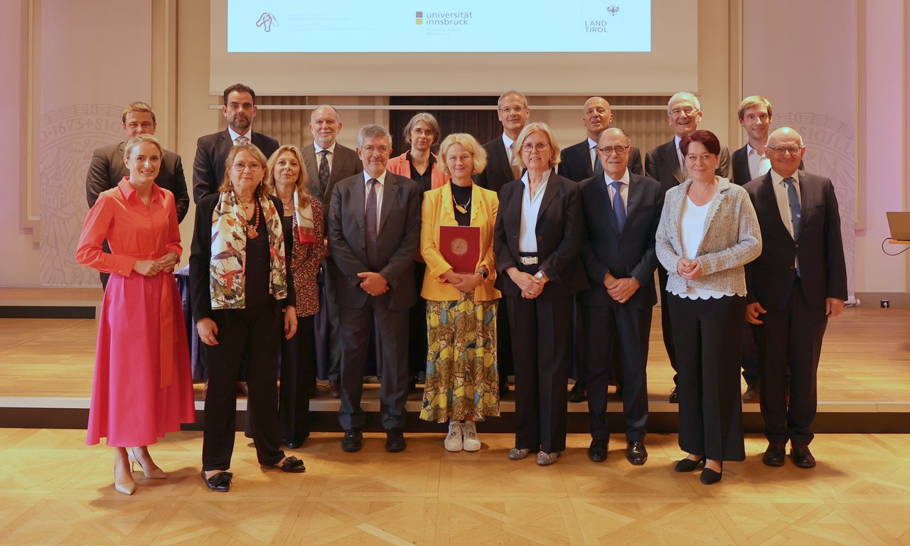 Gruppenfoto von 17 Personen im vorderen Bereich der Aula im Uni-Hauptgebäude.