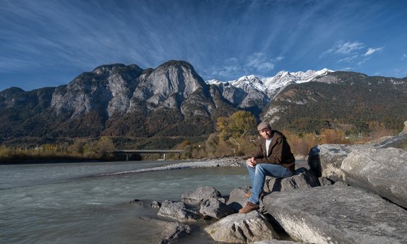 Eine Person sitzt auf einem Stein am Ufer eine Flusses, im Hintergrund schneebedeckte Berge