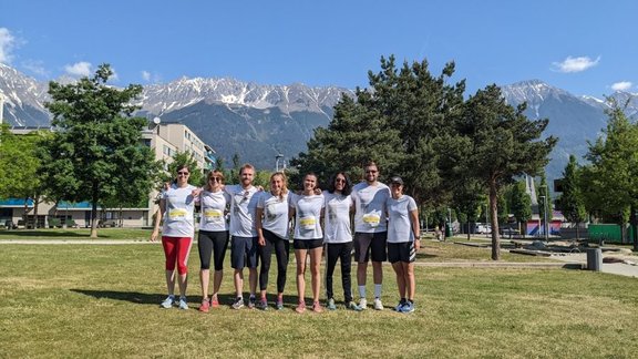 Group photo of eight runners, standing in a line in front of trees and a mountain scenery in the background. All are wearing white university sports shirts.
