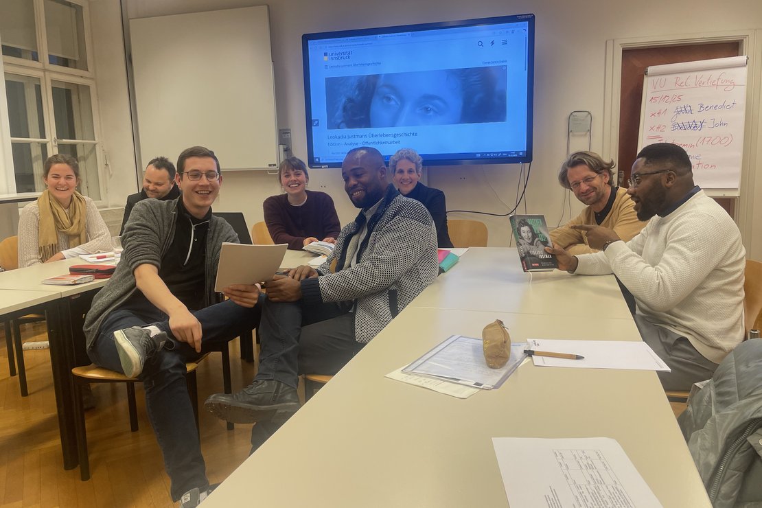 Teilnehmer:innen, LV-Leitung treffen sich mit Dominik Makl und positionieren sich im Sitzen für ein Gruppenfoto. Dabei begutachten sie die vom Projekt erhaltenen Disseminationsprodukte (Grafiknovelle und Übersetzung).