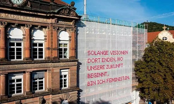 Half-covered building on which is written in german: AS LONG AS VISIONS END WHERE OUR FUTURE BEGINS, I AM FEMINIST:IN.