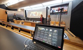 Laptop in einem Hörsaal, auf dem eine Zoom Konferenz zu sehen ist. Im Hintergrund stehen zwei Frauen und ein Mann vor einer Leinwand und halten einen Vortrag.