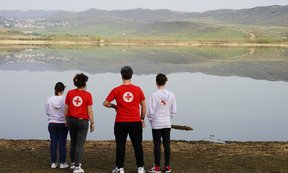 Vier Personen mit T-Shirts des Roten Kreuzes stehen mit dem Rücken zur Kameravor einem See