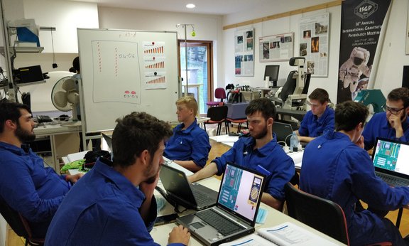 Seven men in blue overalls sit at desks and work on laptops