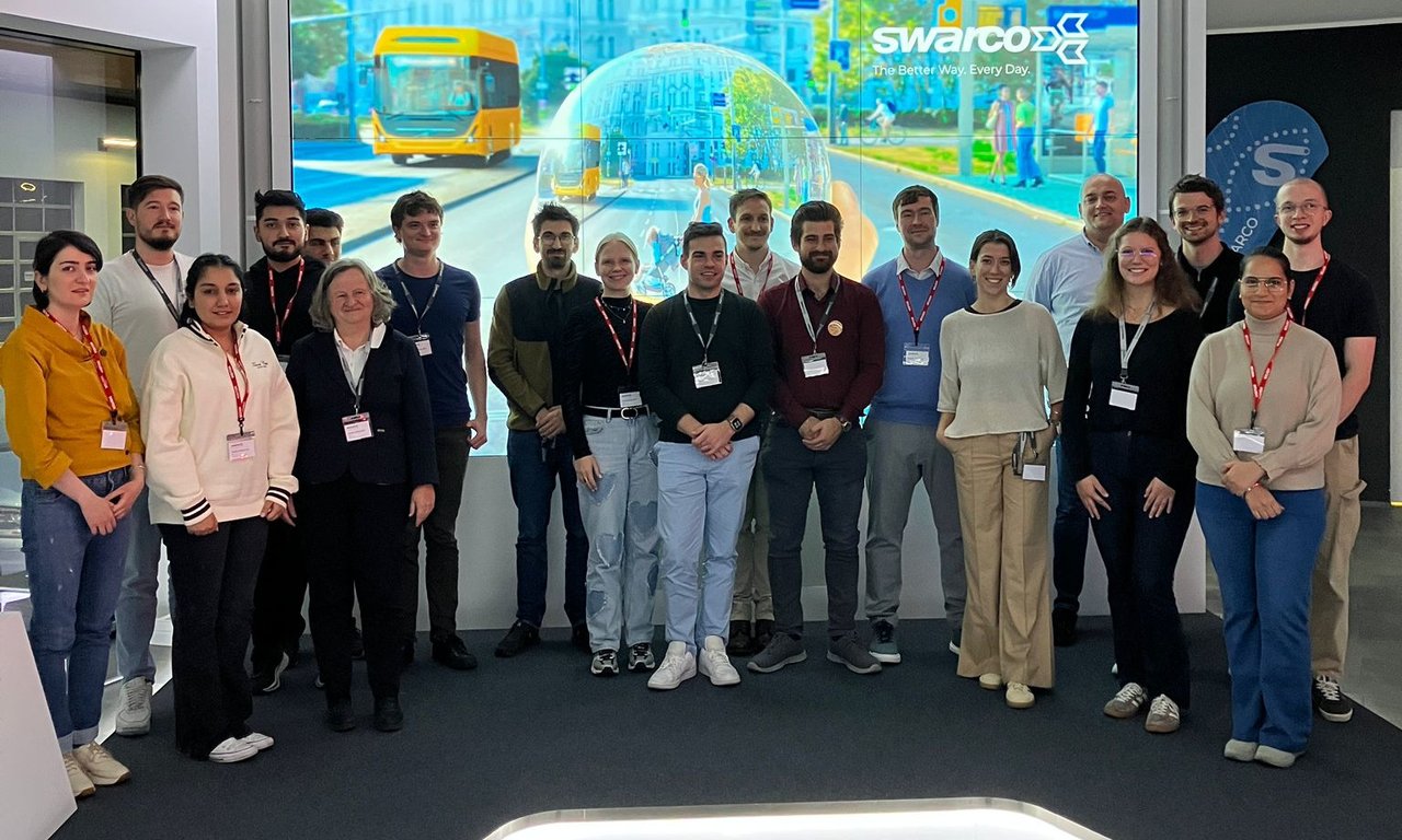Alle Teilnehmer:innen stehen vor einem großen Präsentationsbildschirm, wo man das Swarco Logo sieht