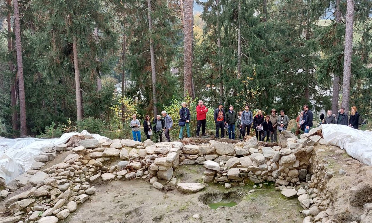 Mehrere Menschen stehen vor ausgegrabenen Steinmauern, diese sind im Vordergrund des Bilds zu sehen