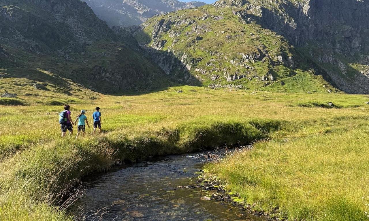 Frau spaziert mit zwei Kindern entlang eines Flusses, im Hintergrund Berge
