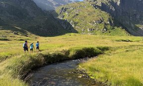 Frau spaziert mit zwei Kindern entlang eines Flusses, im Hintergrund Berge